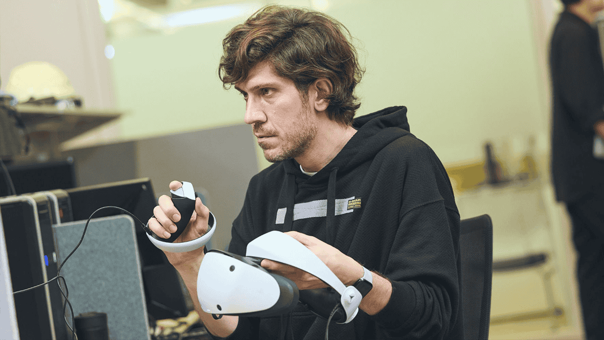 A man wearing a black hoodie holding a PlayStation VR headset in his left hand and a controller in the right hand.